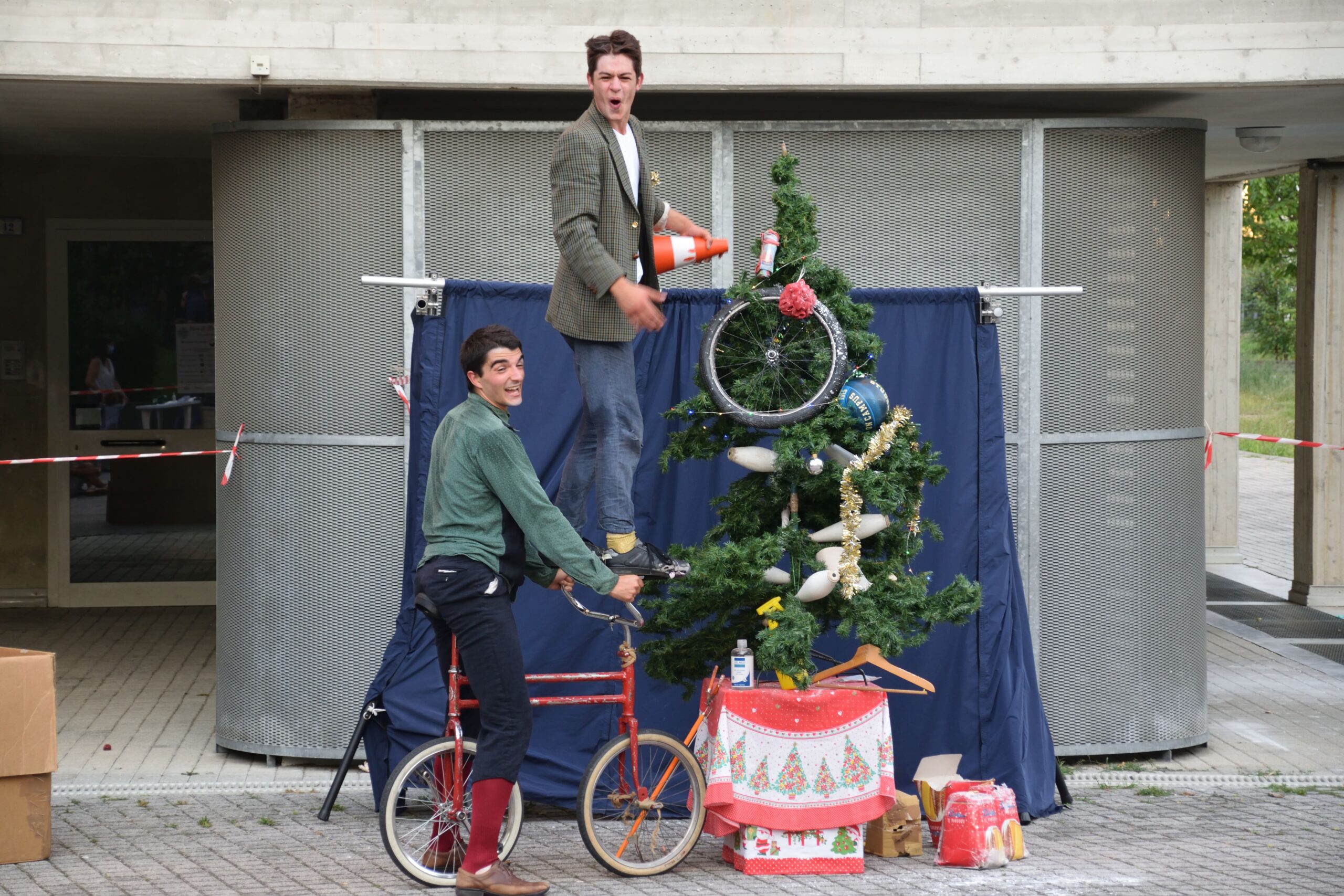 Due clown, uno alla guida di una bici e l'altro in piedi sul manubrio sono fermi davanti ad un albero di Natale decorato con strani oggetti.