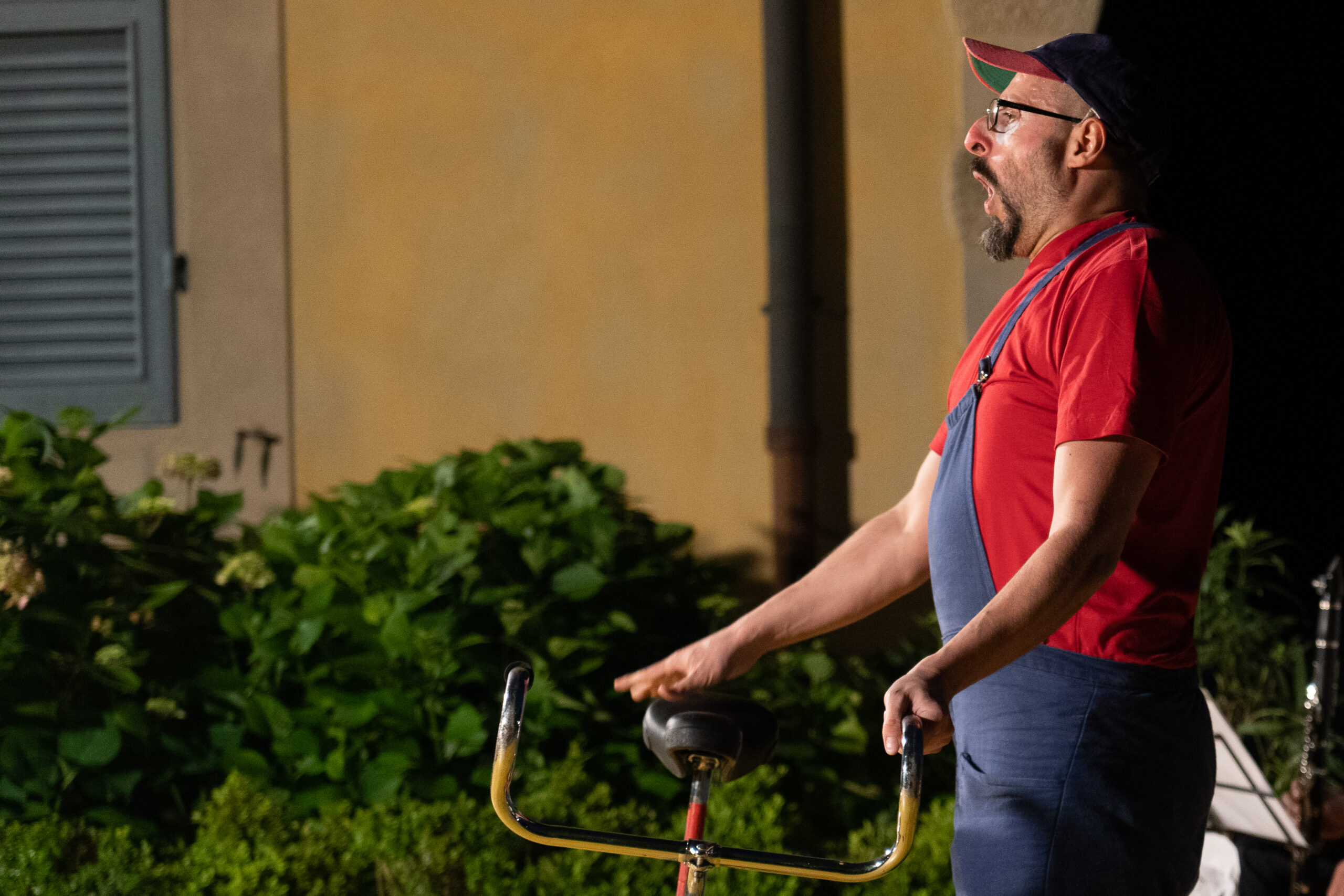 Un clown con una maglia rossa e una salopette blu regge una bicicletta.